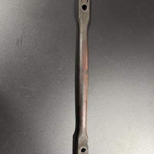 Op de afbeelding: Een donkerbruine, langwerpige metalen handgreep met een gladde, licht gebogen centrale stang. De handgreep heeft twee platte, rechthoekige uiteinden, elk voorzien van een rond gat en een klein, geïntegreerd metalen scharniermechanisme.