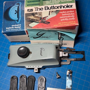 Op de afbeelding: Een vintage Greist knoopsgatenmaker naaimachine-opzetstuk met de originele doos. De doos is blauw en wit met tekst en een diagram. De knoopsgatenmaker is grijs met een zwarte knop en verschillende zwarte sjablonen. De tekst op de doos luidt "The Buttonholer".