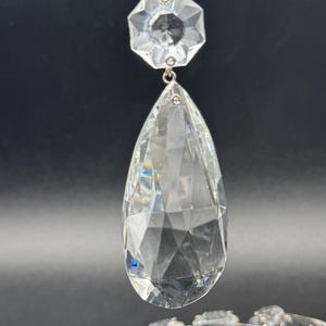 May include: A clear, faceted crystal chandelier pendant featuring a small octagonal top piece connected to a large, elongated teardrop-shaped drop. The crystal is transparent with sharp, geometric cuts that reflect light, suspended by a thin metal wire.