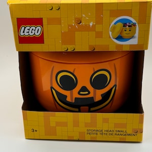 May include: Orange LEGO storage container shaped like a pumpkin with a smiling face. The container is labeled "STORAGE HEAD SMALL" and "PETITE TETE DE RANGEMENT".