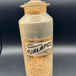 May include: A clear glass apothecary jar with a matching glass stopper, featuring a rectangular label with a gold border and the black text P.JALAP.CO. The jar contains a small amount of light brown, granular material at the bottom.