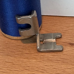 May include: A metal sewing machine foot rests on a wooden surface next to a spool of blue thread. The foot has a U-shaped design with a small bar across the opening. The thread spool is made of wood and has a blue thread wrapped around it.