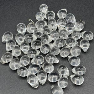 Pode incluir: Uma coleção de contas de vidro transparentes em forma de gota, com um pequeno orifício na parte superior. As contas estão espalhadas sobre uma superfície escura, criando um jogo de luz e sombra. Adequado para fazer joias e projetos de artesanato.