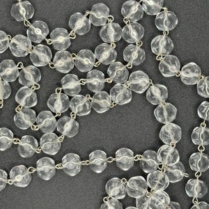May include: A clear, faceted bead necklace with silver-toned metal links. The beads are round with a slightly irregular shape, and the necklace is displayed against a dark background. The beads appear to be made of glass or crystal.