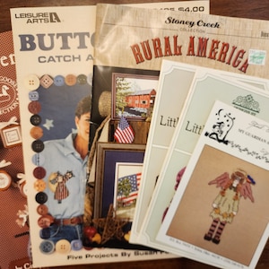 May include: Four cross-stitch patterns featuring a red, white, and blue button theme. The patterns include a country scene with a red barn, a woman with a button-themed dress, a guardian angel, and a small country scene with a red barn. The patterns are from the Stoney Creek Collection, Rural America.