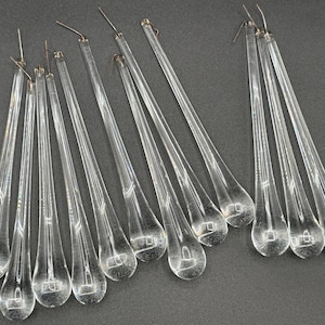 Può includere: Cristalli di lampadario in vetro trasparente a forma di goccia. Ogni cristallo ha un sottile anello di filo metallico nella parte superiore per appenderlo. I cristalli sono disposti su una superficie grigio scuro.