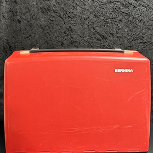 Puede incluir: Un maletín de transporte rígido rectangular de color rojo para máquina de coser. El estuche cuenta con un asa negra en la parte superior y el texto blanco BERNINA impreso en la parte superior derecha. La superficie de plástico rojo tiene arañazos.