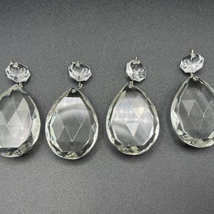 Puede incluir: Cuatro colgantes de cristal transparente para lámpara de araña con forma de lágrima facetada. Cada colgante tiene un cristal facetado más pequeño en la parte superior, conectado por un lazo de metal plateado. Sobre fondo gris oscuro.