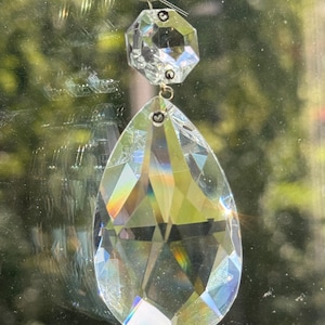 May include: A clear, faceted crystal suncatcher featuring a small hexagonal top piece connected to a larger, teardrop-shaped bottom piece. Both crystal elements are attached to a silver-toned metal ring at the top for hanging purposes.