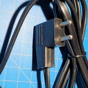 Puede incluir: Primer plano de un cable eléctrico negro con un enchufe de dos clavijas. El enchufe tiene una carcasa de plástico negro y dos clavijas metálicas.
