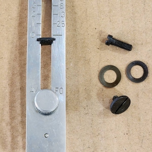 May include: A silver metal sewing machine part with measurements in inches, labeled IN and RE. Includes screws and washers. The part has a dial and a sliding black piece.