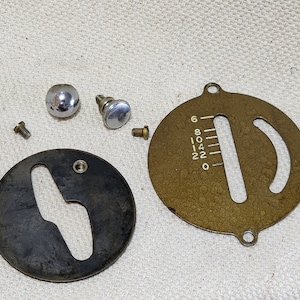 May include: A collection of vintage sewing machine parts, including a brass-colored dial with numbered markings, a black metal cover with a curved slot, several screws, and two chrome knobs. The parts are arranged on a light-colored surface.