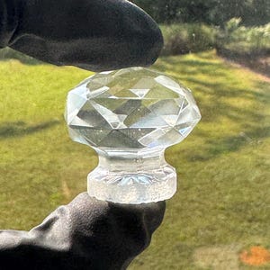 May include: A clear, faceted glass cabinet knob with a diamond-like shape. The knob features a multi-faceted top surface that reflects light, transitioning into a narrower, textured cylindrical base designed for attachment to furniture or cabinetry.