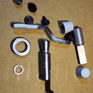 May include: Assortment of sewing machine parts, including a silver metal foot pedal, a white plastic foot, a black plastic knob, a silver metal ring, a white plastic lever, and a silver metal screw.