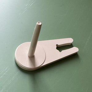 May include: A light beige wooden yarn holder with a vertical dowel and a flat, wing-shaped base. The base has a circular section and a slot for holding yarn. The holder is designed to keep yarn organized during knitting or crocheting.