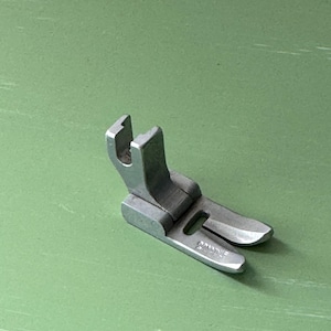 May include: A silver-colored sewing machine presser foot, designed for attaching to a sewing machine. The foot has a flat base with two prongs and a raised, U-shaped section. The item is on a green surface.