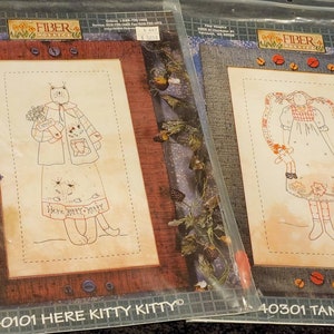 May include: Two embroidery patterns on white paper with a brown border. The pattern on the left is titled "40101 Here Kitty Kitty" and features a woman holding a bouquet of flowers. The pattern on the right is titled "40301 Tansy Angel" and features a woman holding a doll.