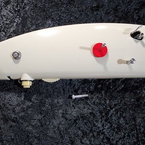 May include: A white sewing machine foot with a red felt pad, a silver screw, and a silver metal hook. The foot is on a black surface.