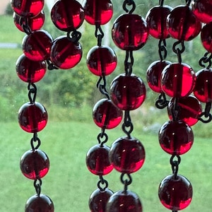 May include: A series of translucent, deep red spherical beads strung together on thin, dark metal links. The beads are arranged in multiple vertical strands, creating a cascading effect where the light shines through the glass-like material.
