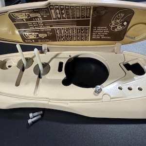 Könnte beinhalten: Eine beige Nähmaschinen-Abdeckung mit geöffnetem Deckel, der eine braune Anleitungstabelle für Stichmuster zeigt. Das Innere verfügt über zwei weiße Kunststoffhebel, eine kreisförmige Öffnung und zwei kleine Metallschrauben an der Basis.