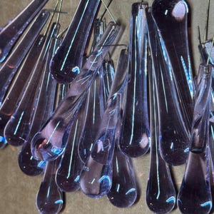 Puede incluir: Primer plano de una colección de adornos de vidrio en forma de lágrima. Los adornos son de color púrpura translúcido con una calidad iridiscente reflectante. Cada lágrima está suspendida por un alambre fino, creando un efecto de araña.