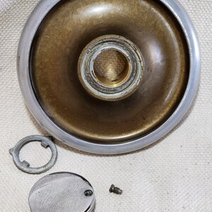 May include: A vintage metal part with a silver rim and a brass-colored center. A small, round, chrome-plated cover with a textured surface, a retaining ring, and a screw are also present.