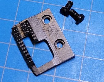 May include: A silver metal sewing machine part with a rectangular shape and a small opening in the center. The part has a series of teeth on one side and the text "SM 47004" on the other side. Two black screws are included.