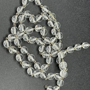 May include: A clear, beaded necklace with round, transparent beads strung on a silver-colored chain. The necklace is laid out on a dark gray surface, showcasing its delicate design and reflective qualities. The beads are evenly spaced, creating a simple yet elegant accessory.