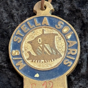 Könnte beinhalten: Vintage Messing-Anhänger mit dem Text "STELLA SOLARIS" in Blau um ein zentrales Schiffsemblem. Der Anhänger zeigt auch die Buchstaben "M/S" und die Nummer "S 12" in Rot.