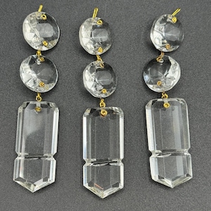 May include: Three clear crystal chandelier pendants. Each pendant has two round crystal beads connected by a gold-colored pin to a rectangular crystal with a pointed bottom. The pendants are displayed against a dark background.