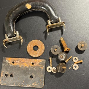 Puede incluir: Un asa de maleta de cuero negro con herrajes metálicos, una placa metálica rectangular con óxido, una arandela metálica circular grande y una colección de varios tornillos, pernos y fijaciones circulares pequeños con signos de oxidación.