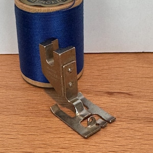 May include: A spool of blue thread and a silver sewing machine foot rest on a wooden surface. The thread spool has a tan cardboard base and a label that reads "STAR SEWING YDS MERCERIZED". The sewing machine foot is made of metal.