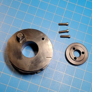 May include: A black metal gear with a small metal knob on top, a smaller black metal ring with three holes, and three small silver screws. The items are on a blue grid background.