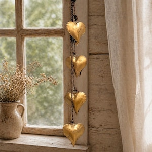 May include: A decorative hanging ornament featuring four gold-colored heart-shaped bells suspended from a rope. The hearts have a textured surface and are hanging in front of a window with a neutral-colored curtain.