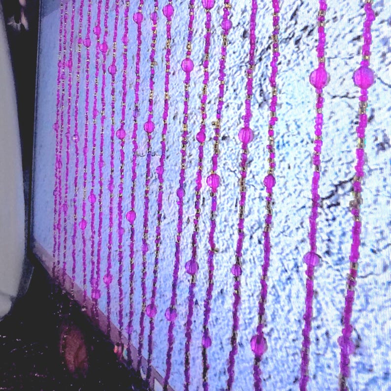 May include: A beaded curtain with strands of small, gold-colored beads and larger, translucent magenta beads. The curtain hangs in front of a textured, light blue wall.