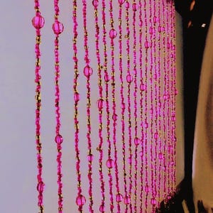 May include: A beaded curtain with strands of pink and gold beads. The pink beads are larger and more rounded, interspersed with smaller gold beads. The curtain hangs in vertical rows, creating a textured and decorative effect.