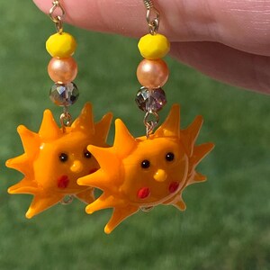 May include: A pair of dangle earrings featuring a bright orange sun charm with a smiling face, red cheeks, and black eyes. Above the sun charm, each earring has a faceted yellow bead, a smooth peach-colored pearl, and a dark iridescent crystal bead.