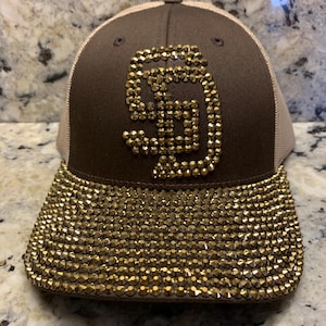 May include: A brown baseball cap featuring a mesh back and a flat brim. The front panel displays the letters SD in gold-toned rhinestones, while the entire brim is densely covered in matching gold-toned rhinestones, creating a textured, reflective surface.