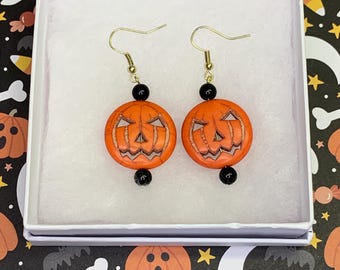 May include: A pair of dangle earrings featuring round, orange, carved jack-o'-lantern beads. Each earring includes a small, round, black bead above and below the pumpkin, suspended from a gold-toned metal hook wire in a white gift box.