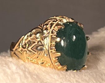 May include: A gold ring featuring an oval-shaped, dark green cabochon gemstone set in a prong setting. The band and the setting are crafted with intricate, raised floral and leaf-like engravings that cover the entire outer surface of the metal.