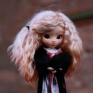 Può includere: Una bambola con lunghi capelli ricci rosa chiaro, che indossa un cappotto di velluto nero con un nastro rosa e un vestito di pizzo bianco. La bambola è in piedi su un piedistallo bianco.