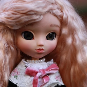 Può includere: Una bambola con lunghi capelli ondulati rosa chiaro, che indossa un cappotto di velluto nero con un colletto di pizzo bianco e un fiocco di raso rosa. La bambola ha gli occhi azzurri e indossa un vestito bianco con pizzo.