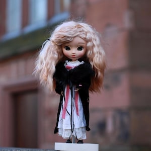 Può includere: Una bambola con lunghi capelli biondi ricci che indossa un cappotto nero con un vestito bianco sotto. La bambola indossa un nastro rosa intorno al collo e un vestito a motivi bianco e nero.