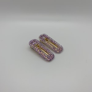Puede incluir: Dos pinzas para el pelo transparentes con purpurina morada incrustada en el plástico. Las pinzas son de forma rectangular y tienen un resorte de metal dorado.