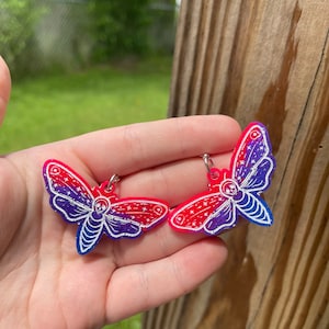 May include: A pair of dangle earrings featuring a moth design with a gradient of pink and purple colors. The moth has a skull and crossbones design on its body and is covered in glitter.