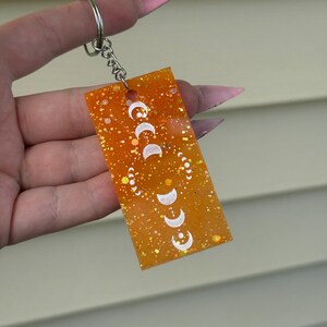 May include: Orange resin keychain with a silver key ring and a white moon phase design.