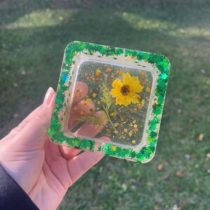 May include: Square clear resin tray with a green glitter border and a pressed yellow flower inside. The tray is filled with gold glitter.