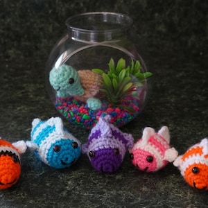 May include: A clear glass fishbowl with a crocheted turtle and a small plant inside. Five crocheted fish in various colors, including orange, blue, purple, and pink, are arranged in front of the bowl. The fish have white fins and smiling faces.