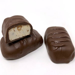 Op de afbeelding: Twee rechthoekige chocoladesnoepjes met een gladde donkerbruine laag. E&eacute;n snoepje is doormidden gesneden om een romige, gebroken witte vulling te onthullen met kleine, zichtbare textuurdeeltjes die in het midden van het snoepje zitten.