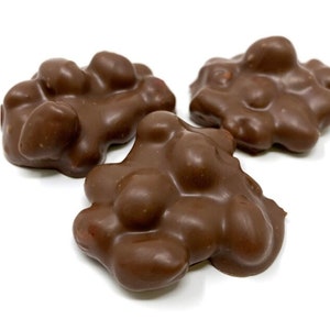 Op de afbeelding: Drie clusters van met melkchocolade omhulde pinda's. Elk stuk heeft een hobbelige, onregelmatige oppervlaktestructuur die wordt gecre&euml;erd door de afgeronde vormen van de noten, bedekt met een gladde, glanzende bruine chocoladelaag.